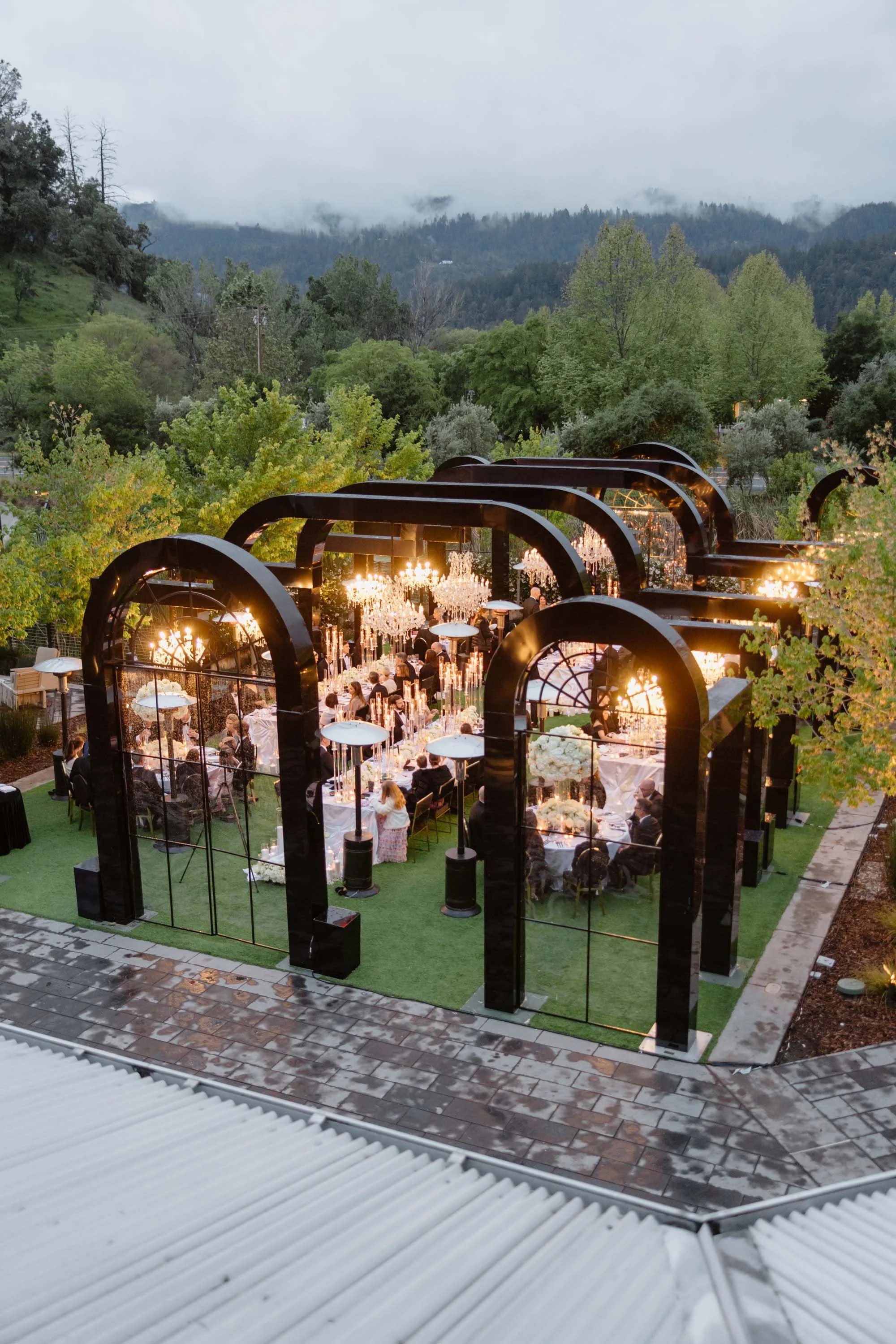 An outdoor evening event with a decorated tent and chandeliers, set in a lush green landscape with trees and mountains in the background.