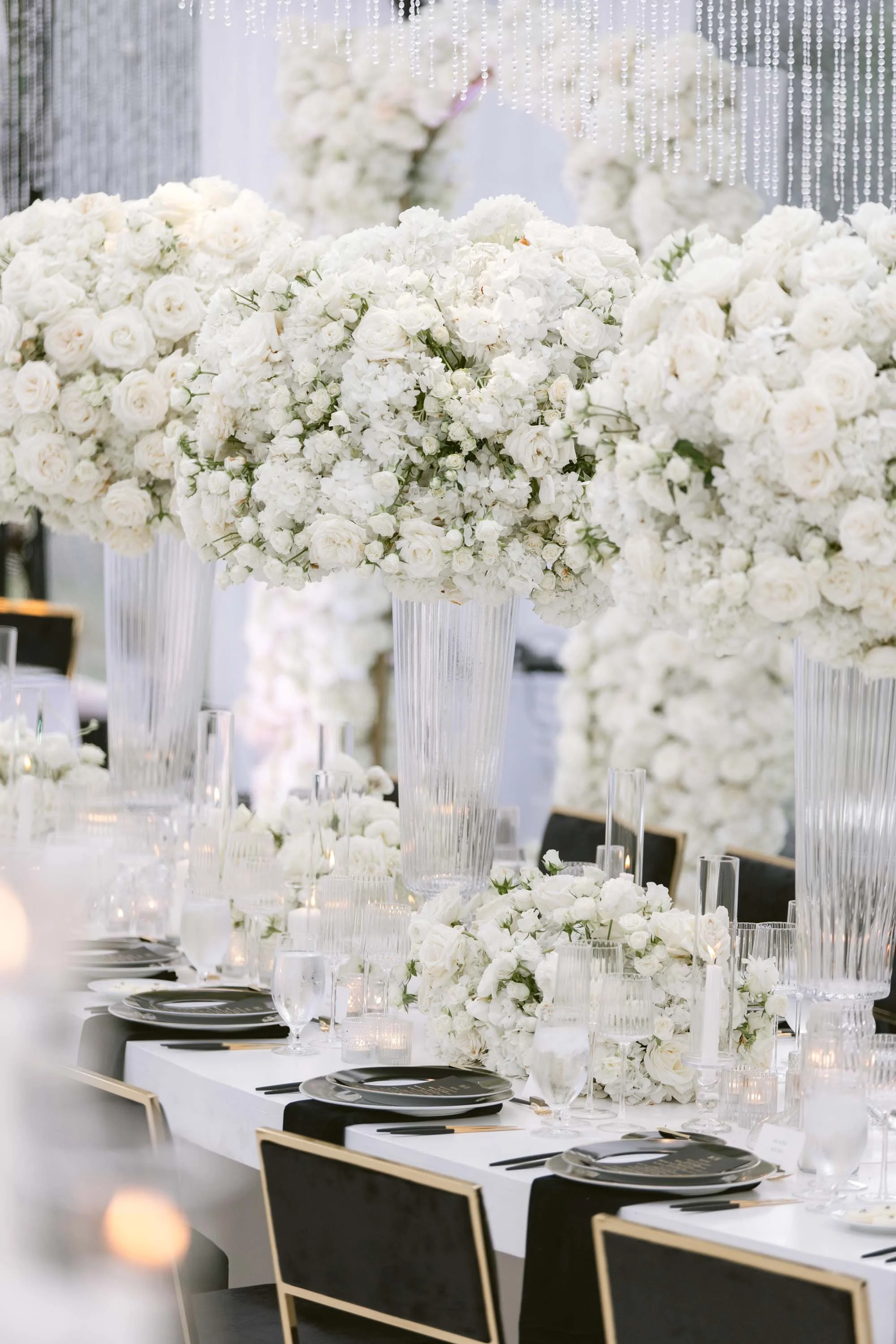 Elegant banquet table decorated with large white floral arrangements in clear glass vases, surrounded by candles, black and gold plates, and clear glassware, set for a wedding or formal event.