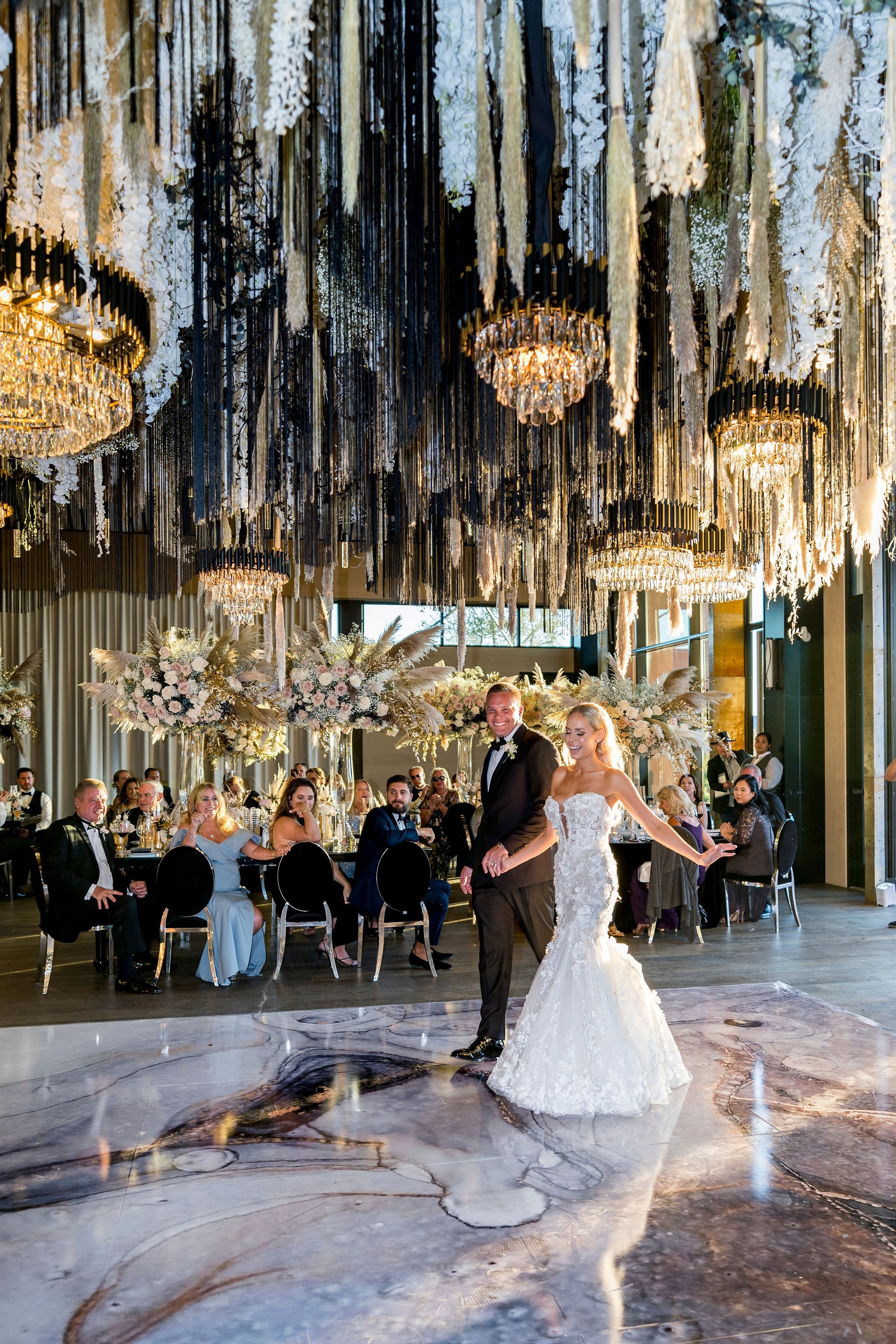 A bride and groom are dancing at their wedding reception in a decorated banquet hall with floral arrangements and chandeliers hanging from the ceiling.