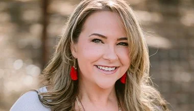 A woman smiling outdoors, wearing red earrings and a light-colored top.