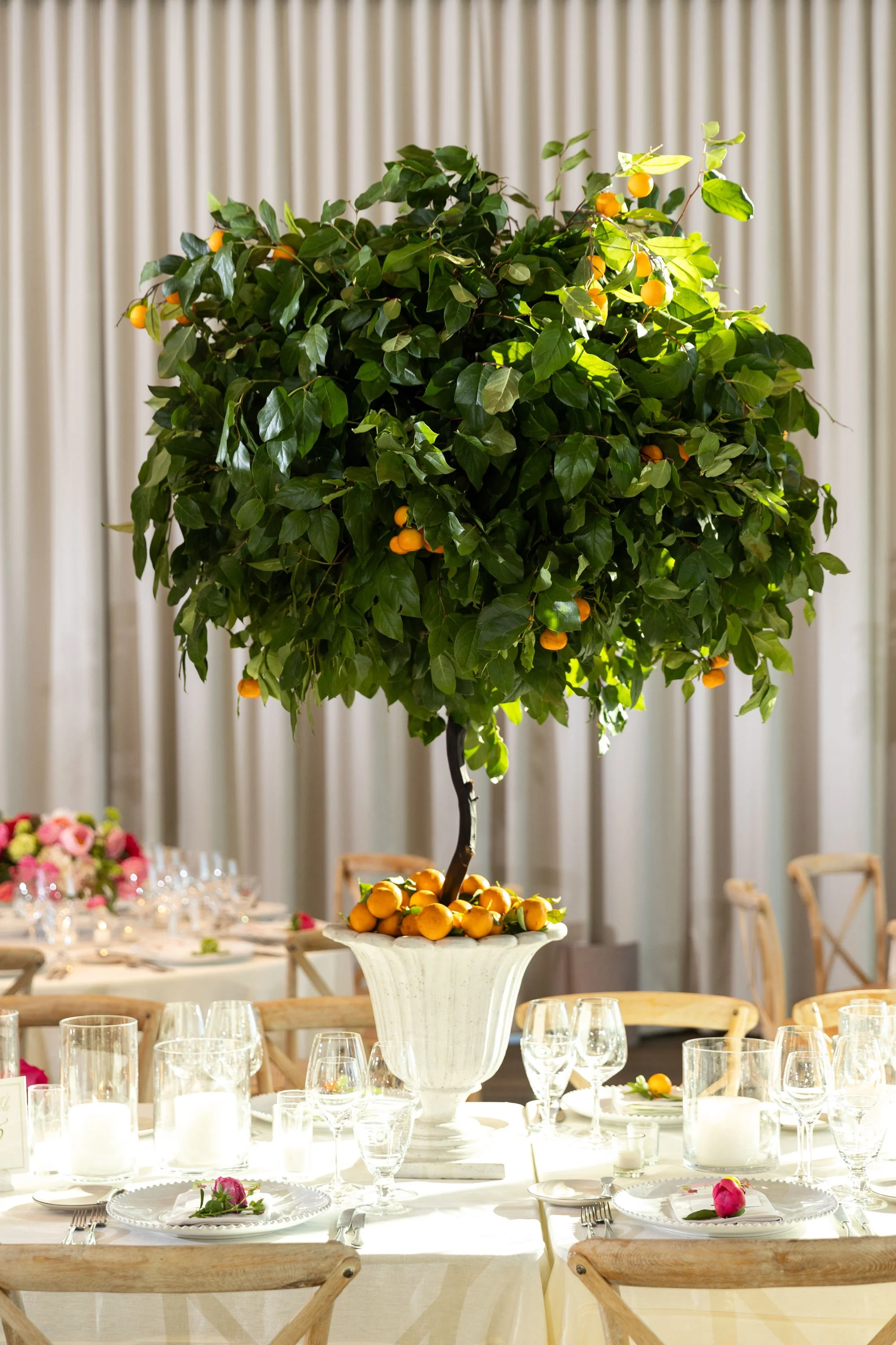 Elegant dining table setup with a large ornamental tree in the center. The table is decorated with wine glasses, candles, and pink roses, with chairs around it. The background features a curtain.