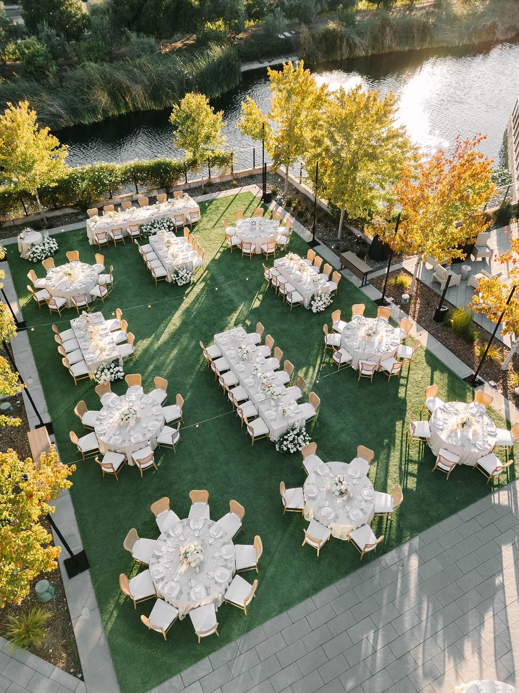 Outdoor event space with round and rectangular tables with white tablecloths, set with plates and glasses, surrounded by beige chairs on green artificial grass, next to trees with yellow and orange leaves, and a body of water in the background.