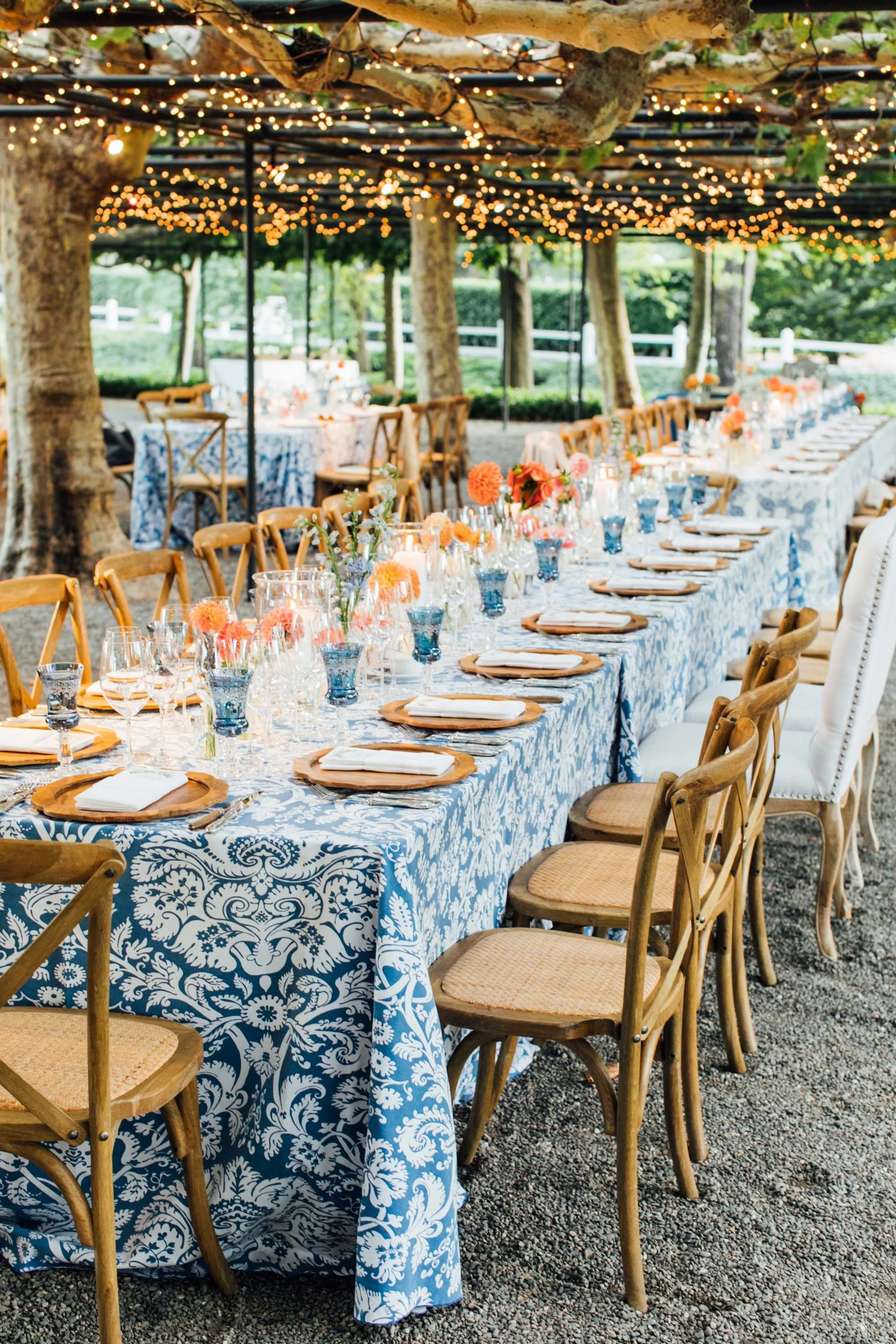 Long dining table set outdoors under string lights, with floral centerpieces, wooden chairs, and place settings, surrounded by trees and greenery on a gravel ground.