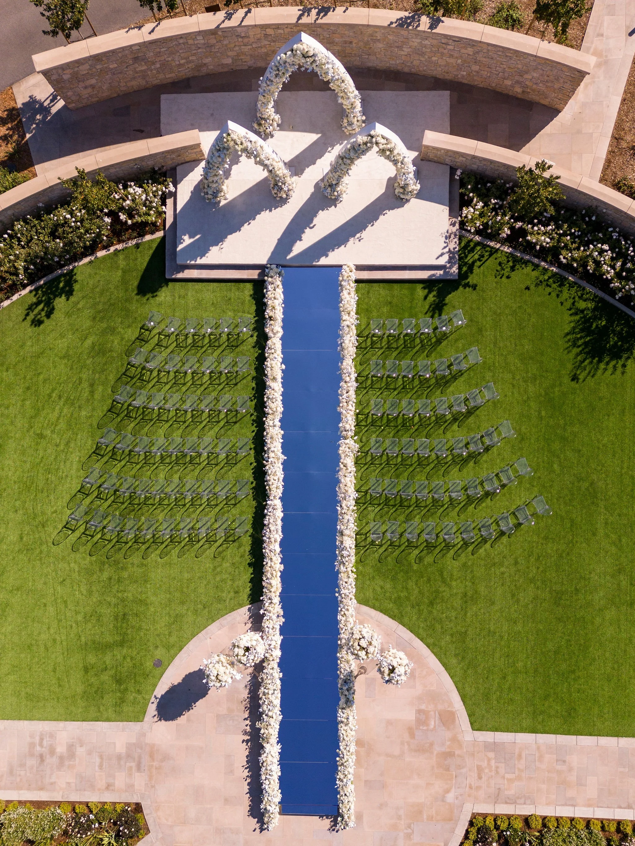 Aerial view of a wedding ceremony setup with a white floral aisle runner, an arch decorated with white flowers, and rows of clear chairs arranged on green grass.
