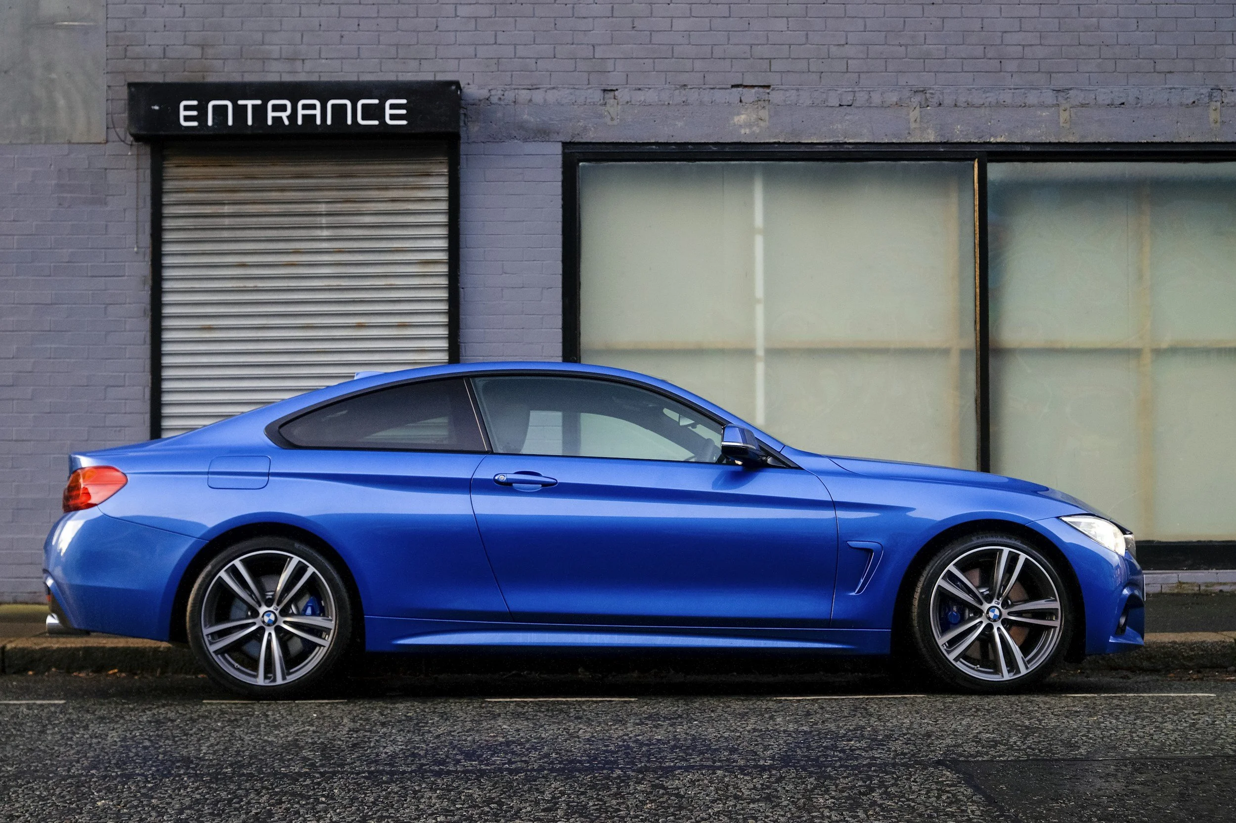A blue sports car parked in front of a building with a sign that says 'ENTRANCE', a brick wall, and a large window.