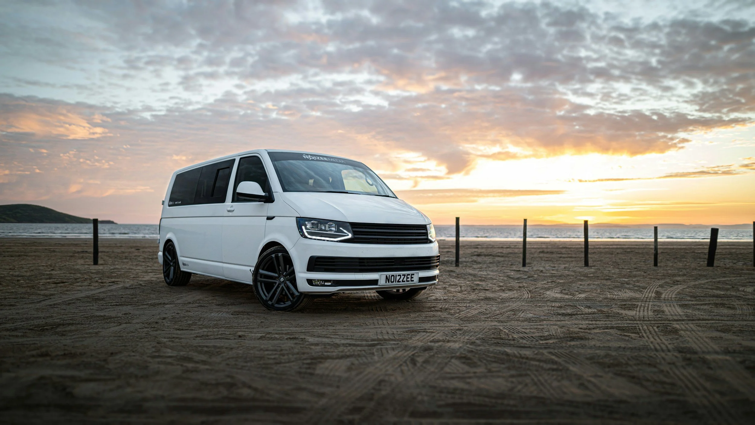 A white van with custom black rims parked on a sandy beach during sunset, with a cloudy sky and ocean in the background.