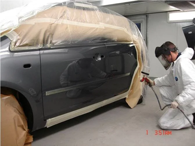 A person in protective gear spray-paints a car in a workshop.