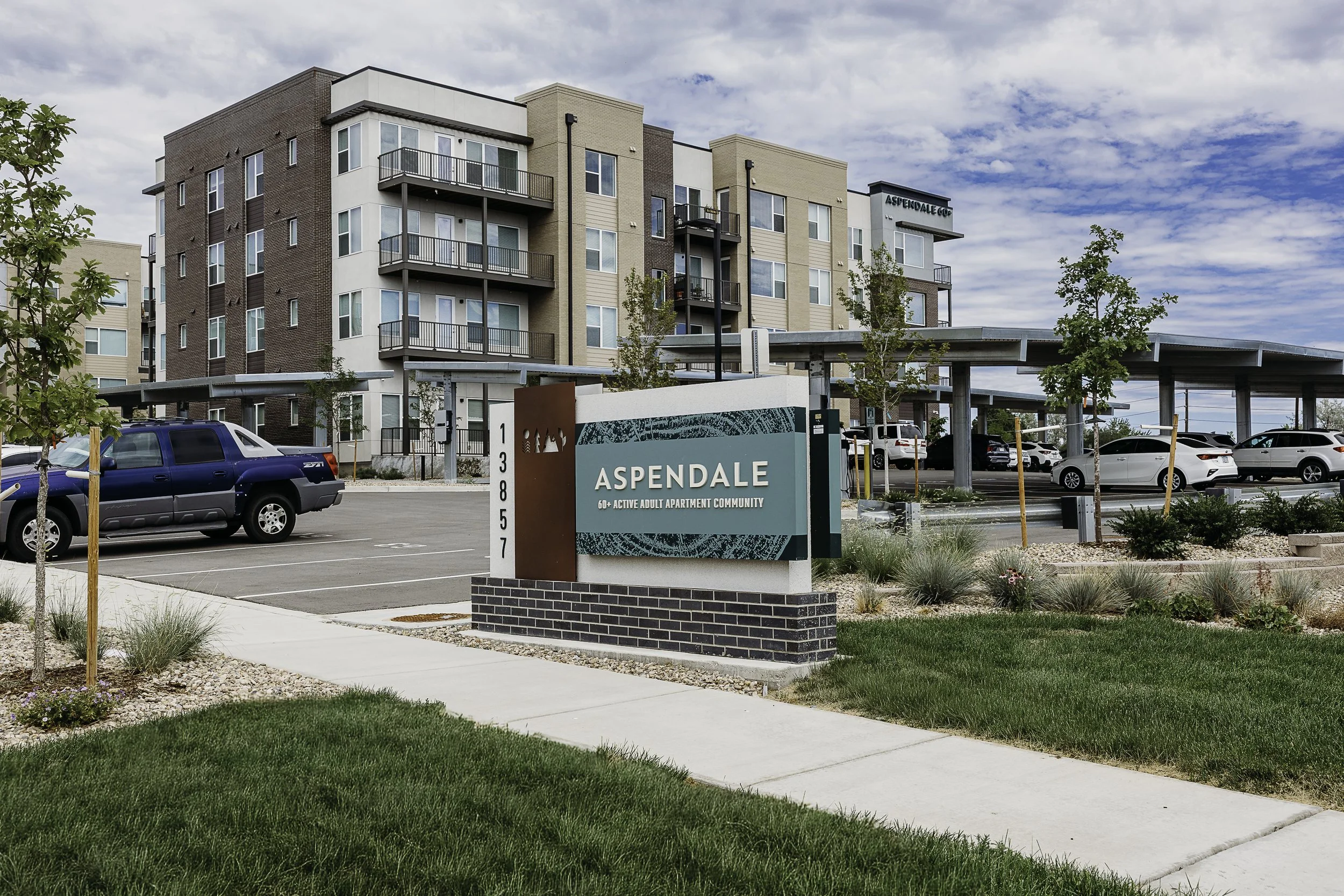 Modern apartment complex named Aspendale with multiple floors, balconies, and parking lot, under a partly cloudy sky.