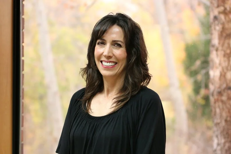 A woman with dark wavy hair and a big smile standing in front of a window with autumn trees outside.