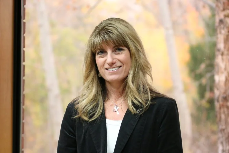 Woman with shoulder-length blond hair smiling, standing in front of a nature background with trees and fall foliage.