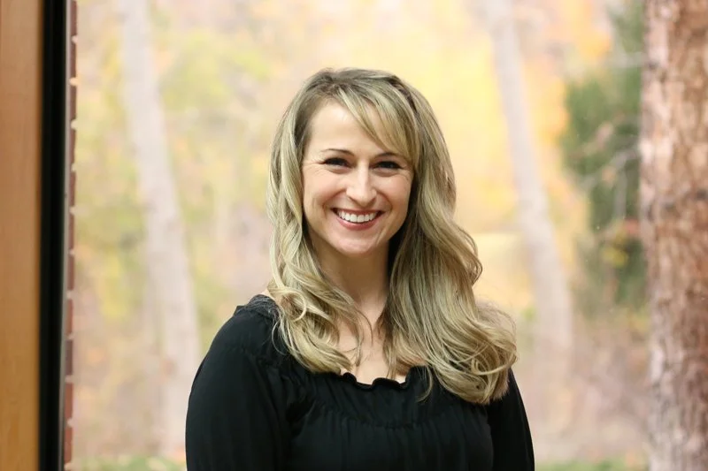 A smiling woman with blonde hair, wearing a black top, standing outdoors in front of a blurred background of trees and autumn foliage.