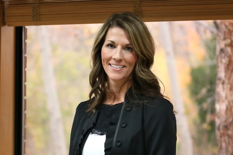 A smiling woman with shoulder-length brown hair, wearing a black jacket and a black top with lace detail, standing indoors near a window with a blurred autumn forest background.