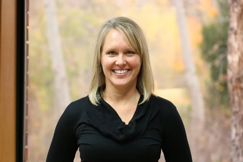 A woman with blonde hair and a black top smiling outdoors with trees and autumn foliage in the background.