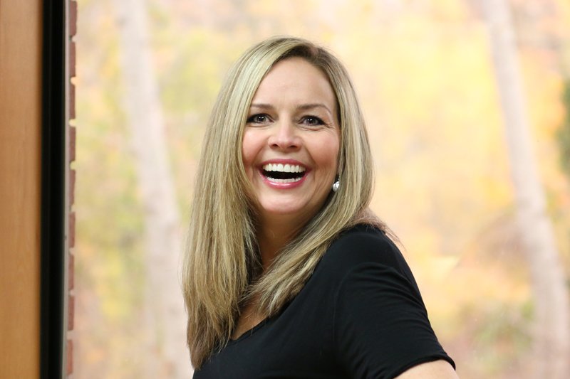 Woman with long blonde hair smiling and looking at the camera, standing indoors near a window with a blurred outdoor background.
