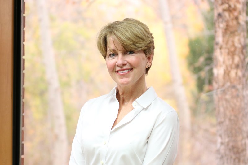 A woman with short light brown hair, wearing a white blouse, smiling and standing outdoors with a blurred forest background.