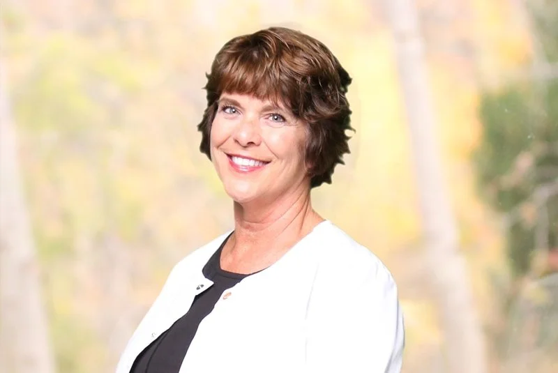 Portrait of a smiling middle-aged woman with short brown hair, wearing a white jacket and black top, standing outdoors with blurred trees in the background.