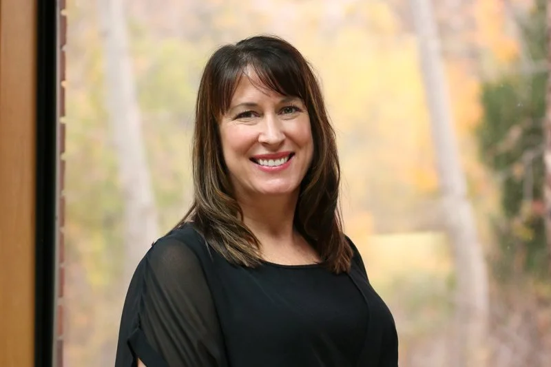 A woman with shoulder-length brown hair and a black top smiling in front of a window with a colorful autumn forest background.