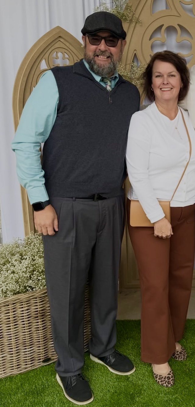 A man and a woman standing together indoors. The man is wearing a dark cap, glasses, a teal shirt, a black vest, and gray pants. The woman is wearing a white top, brown pants, and leopard print shoes, holding a tan purse. Both are smiling, with a decorative background and green grass-like carpet on the floor.