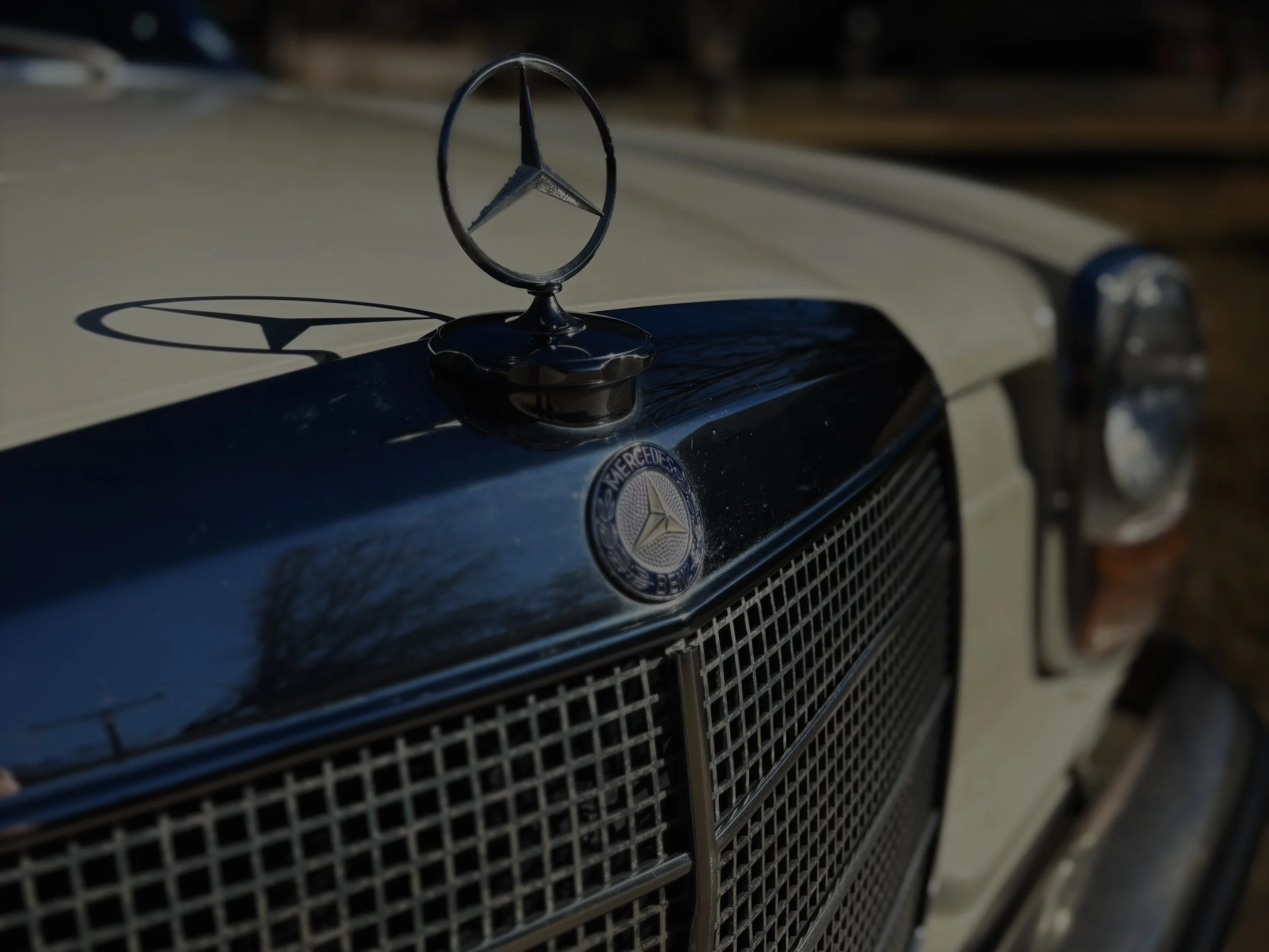 Close-up of a Mercedes-Benz car hood ornament with a shadow cast on the hood, and a metal grille with the Mercedes-Benz emblem visible below.