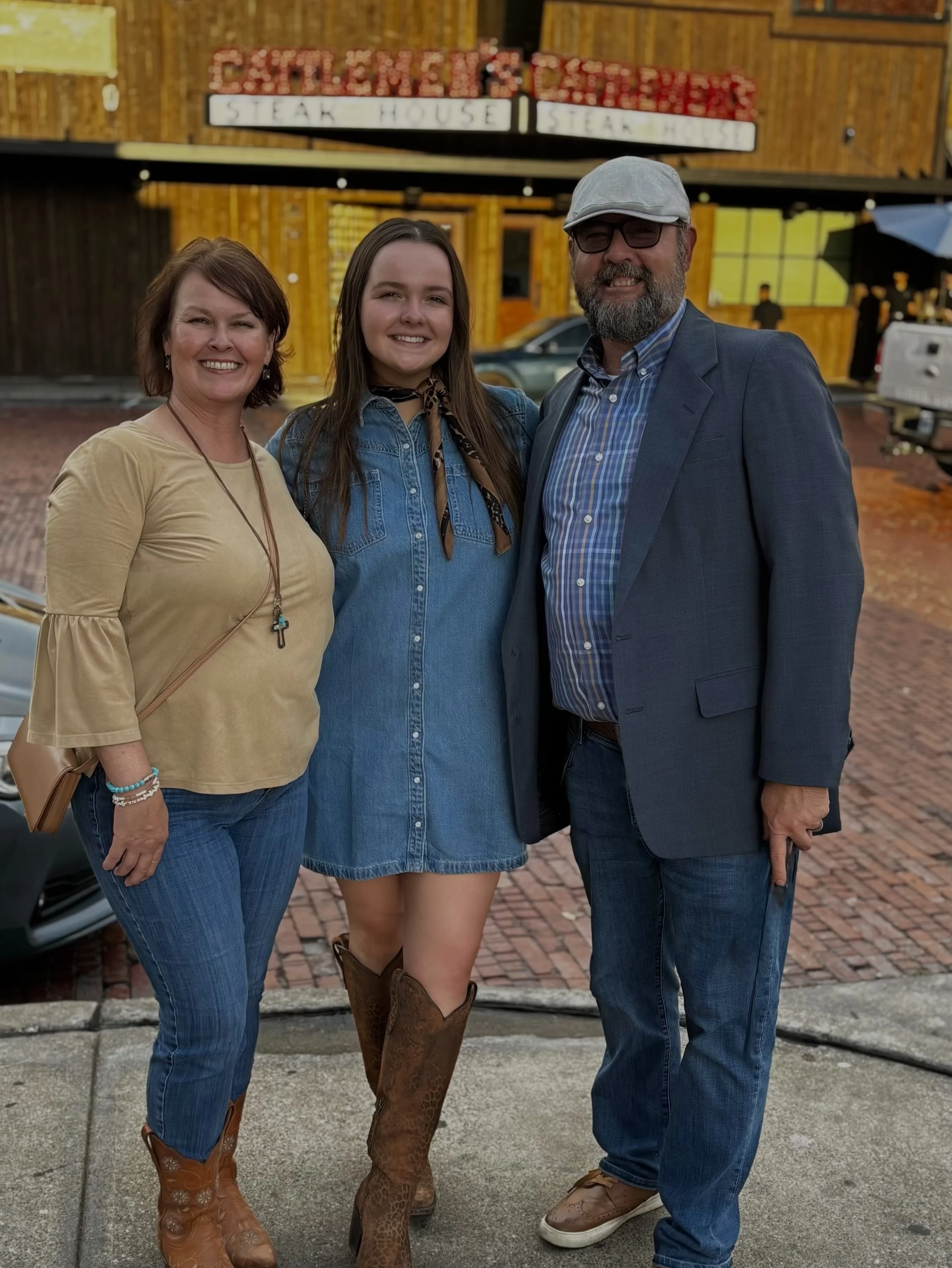 Three people standing outside a restaurant called 'CATERING & STEAKHOUSE' in front of a yellow building, smiling at the camera. The woman on the left has short brown hair, wearing a beige top, blue jeans, and cowboy boots. The young woman in the middle has long dark hair, wearing a denim dress, a bandana, and cowboy boots. The man on the right has a beard, glasses, a gray cap, a blue blazer, and casual shoes.