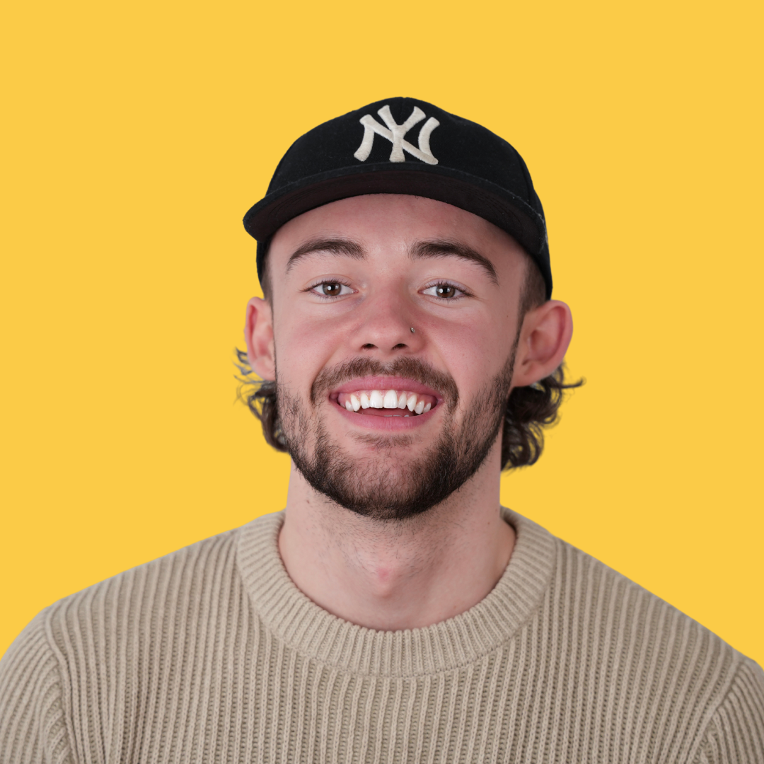 Un jeune homme souriant, portant une casquette noire avec le logo des New York Yankees, un pull beige, et un petit nez percé, contre un mur jaune.