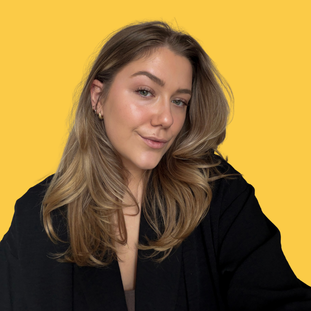 Une femme aux cheveux longs, blonds et ondulés, portant une veste noire, sourit légèrement devant un fond jaune.