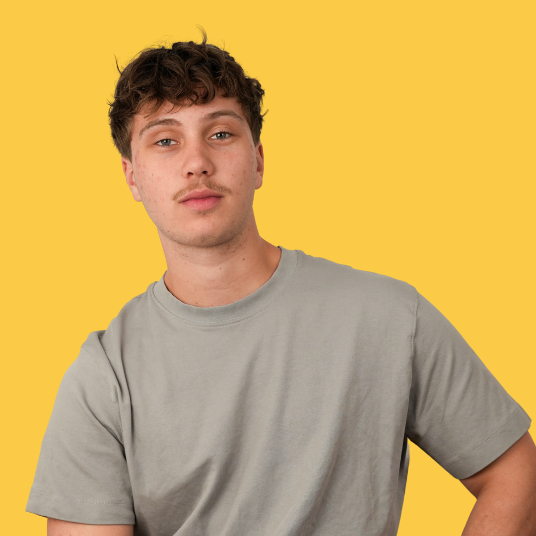 Jeune homme avec cheveux bruns bouclés et peau claire, portant un t-shirt gris, souriant légèrement, face à un fond jaune.