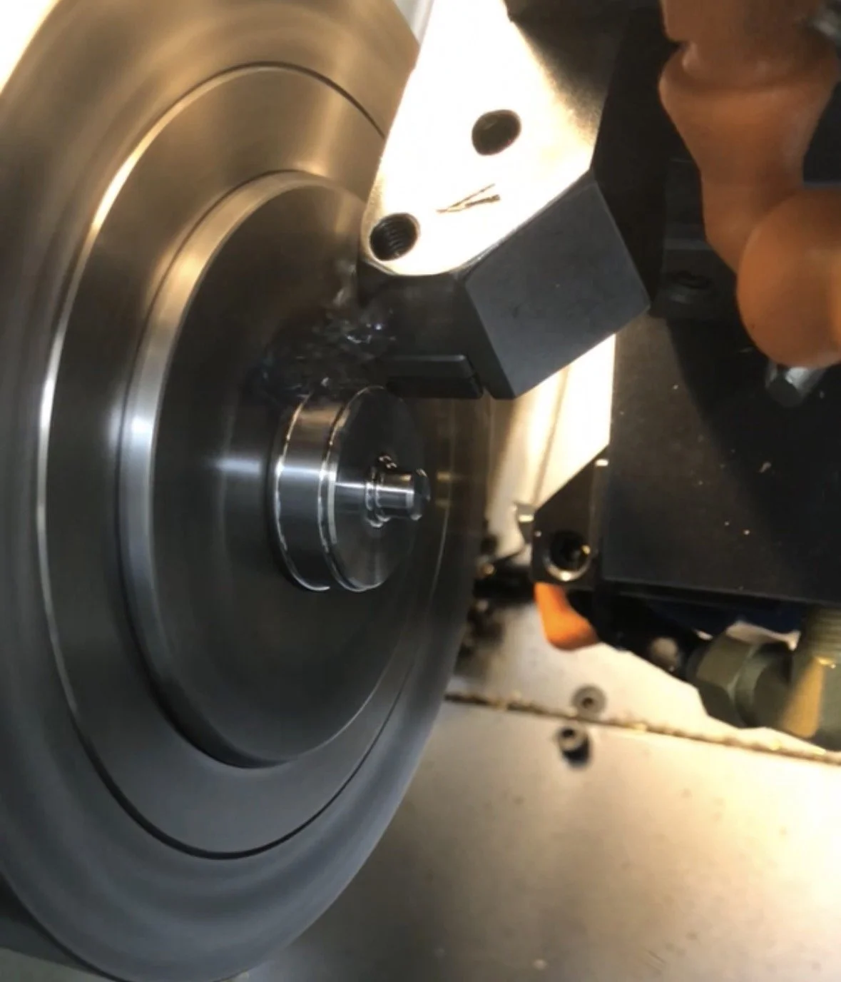 Close-up of a metal lathe machine with a rotating cylindrical piece being machined, and a person with gloves operating the equipment.