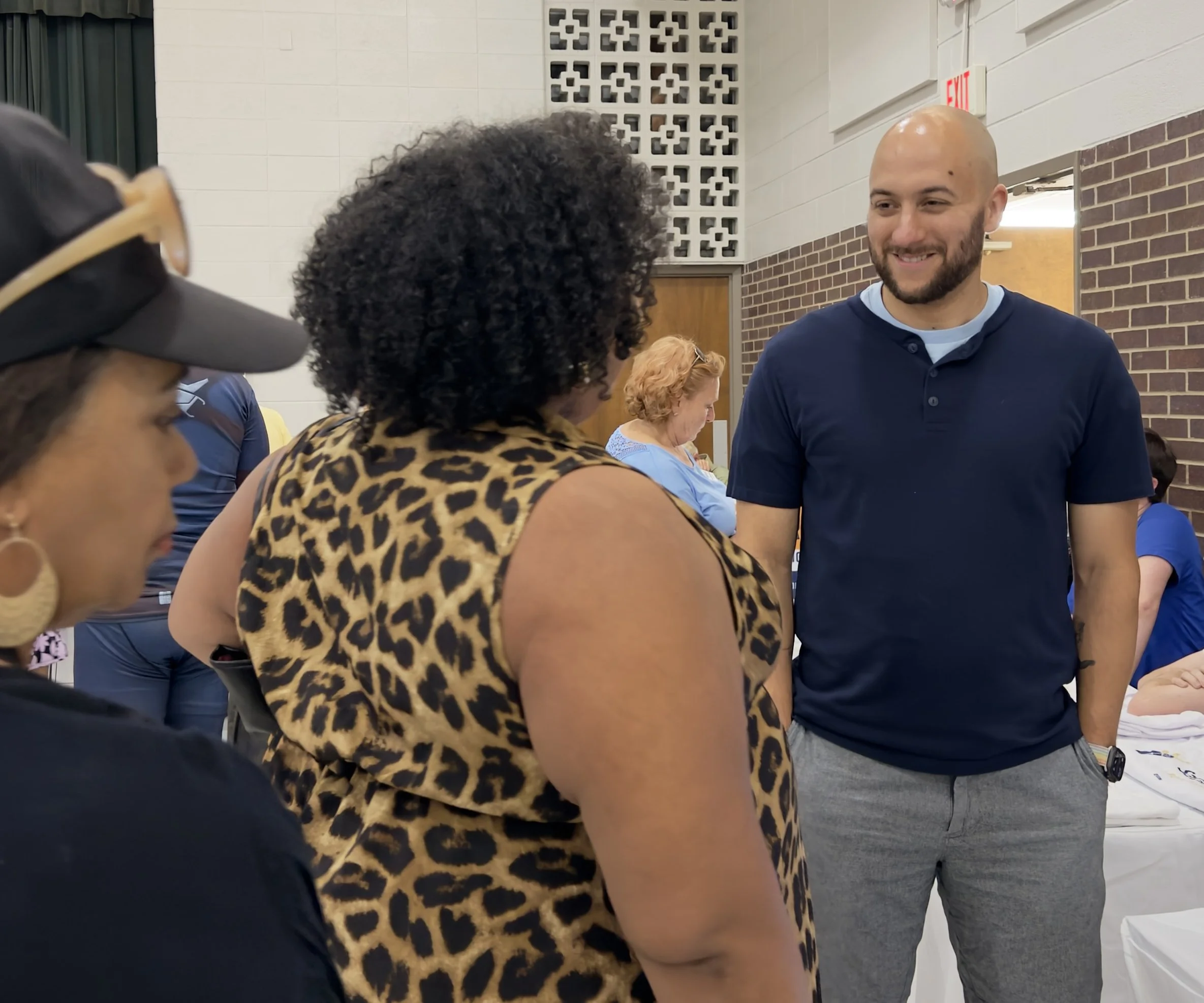 A group of people in a room talking, including a man with a bald head and beard smiling, facing a woman with curly black hair and a leopard print top, while others are partially visible around them.