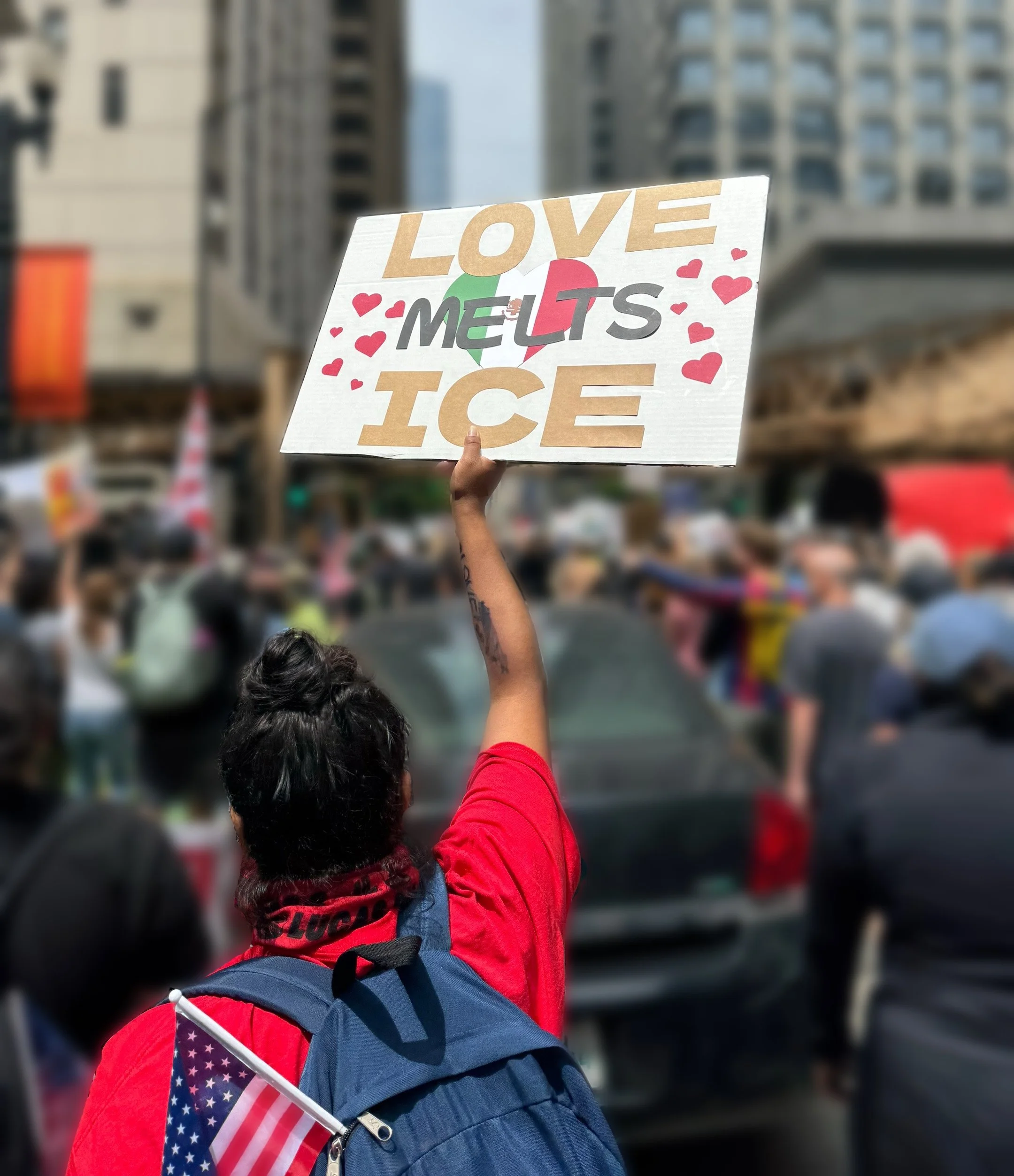 A person wearing a red shirt with a backpack holding a sign that reads 'Love Melts Ice' with the 'O' in 'Love' represented by a heart-shaped diagram with American flag colors. The person is in a crowd on a city street with tall buildings in the background.
