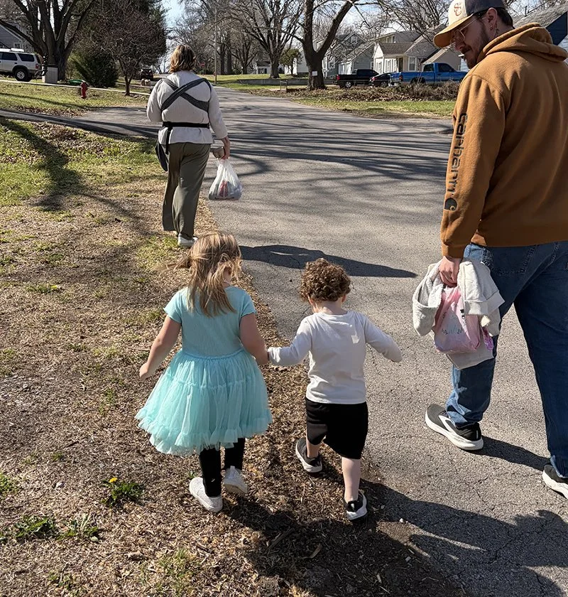 Two young children, a girl in a blue dress and a boy in a white hoodie, walk hand-in-hand along a sidewalk with an adult man nearby, in a neighborhood on a sunny day.