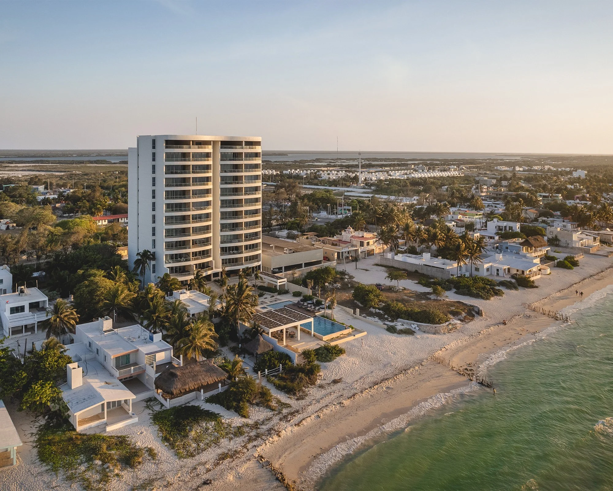 Vista aérea de la playa con arena clara y olas suaves, casas y hoteles blancos y cabañas con techos de paja, palmeras, y un edificio alto de forma circular en primer plano, al atardecer o amanecer en una zona costera tropical.