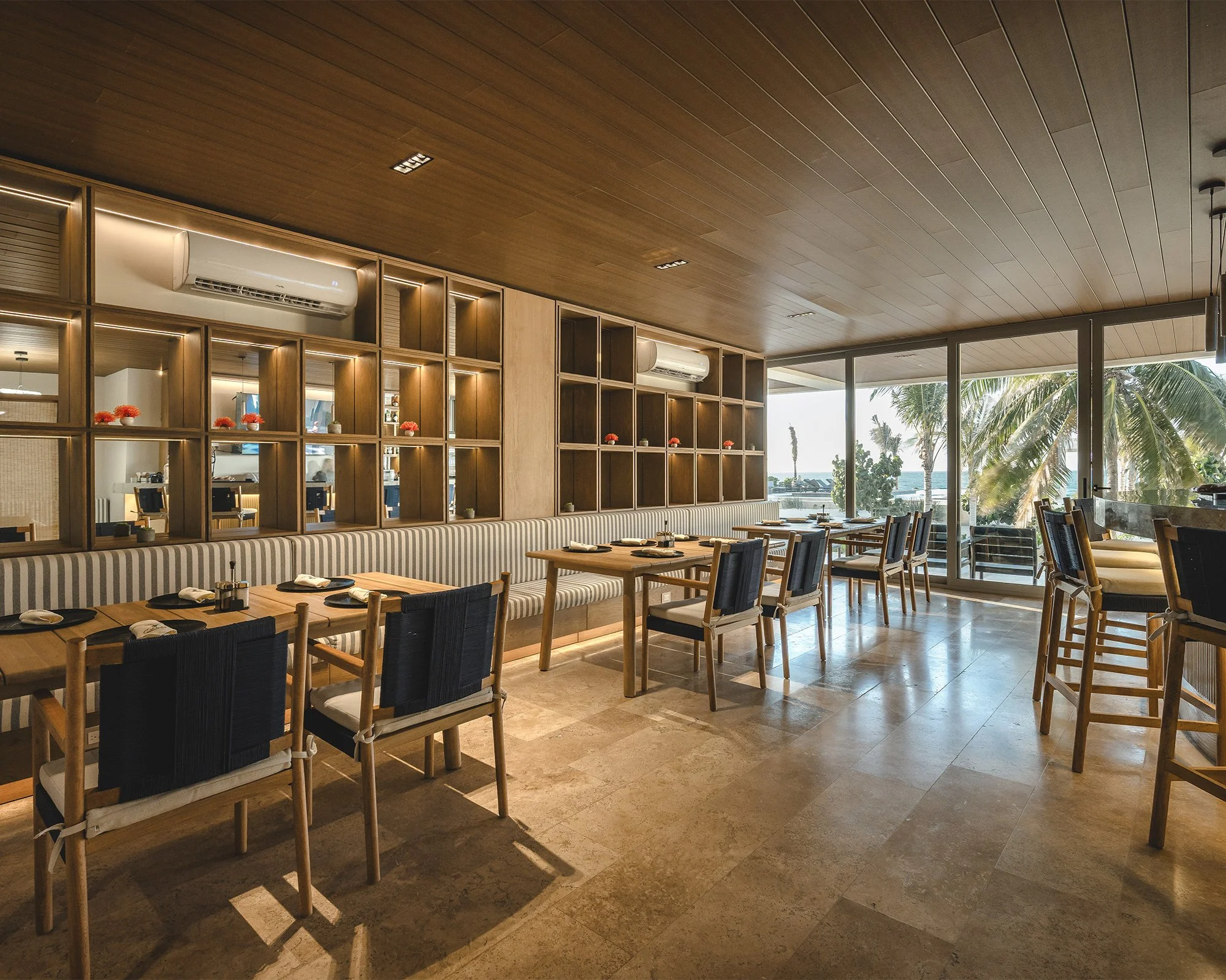 Interior de un restaurante con mesas de madera, sillas de madera y tela, estantes de madera con decoraciones, y ventanas grandes que muestran palmeras y vista al mar.