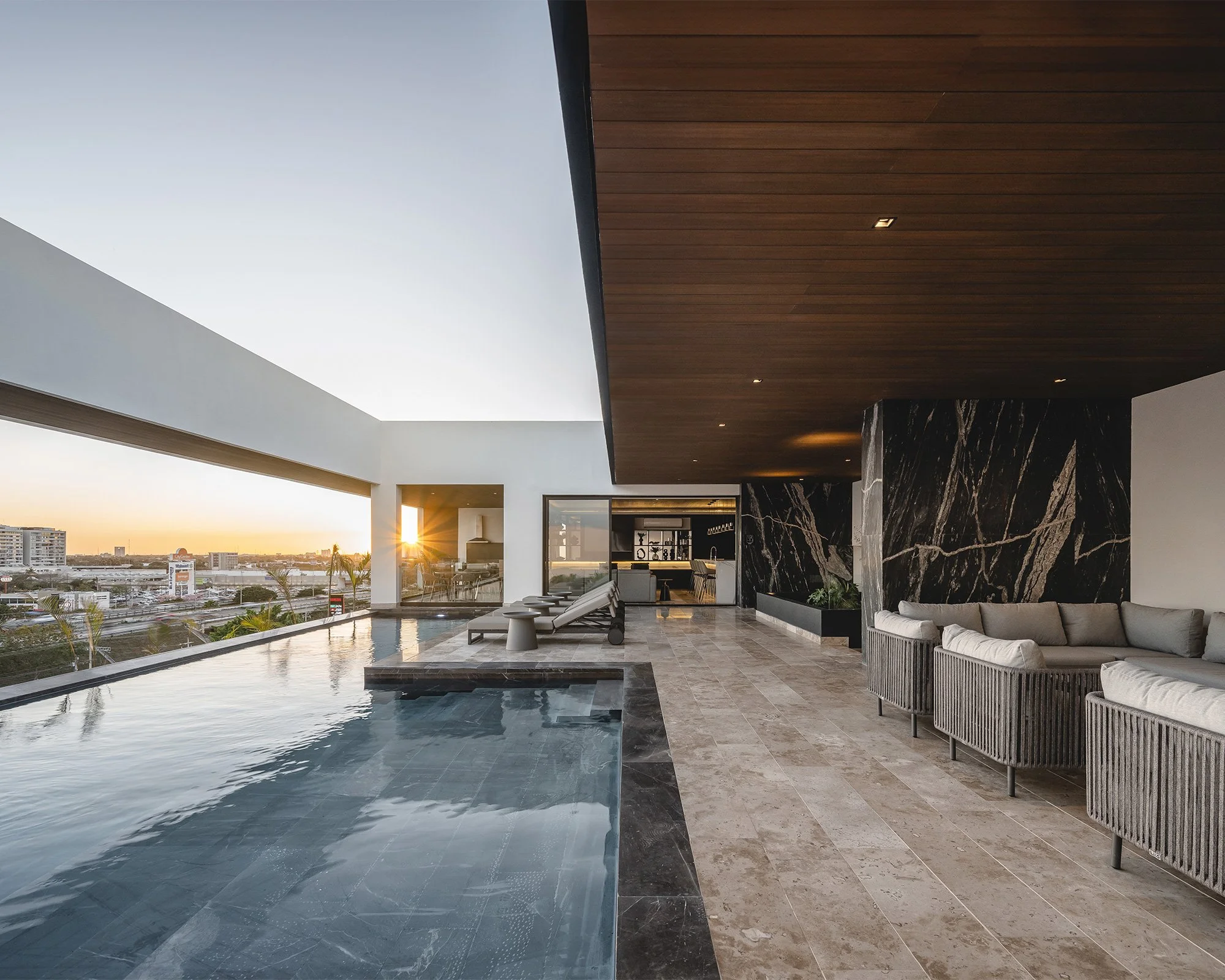 Vista de una terraza moderna con piscina, muebles de exterior y una vista del atardecer en la ciudad, bajo un techo de madera.