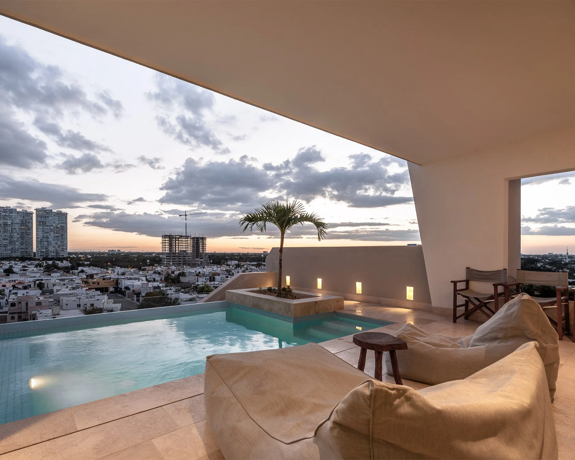 Vista de una terraza en la azotea con una piscina y sillones, con la ciudad y edificios altos de fondo durante el atardecer, cielo con nubes.