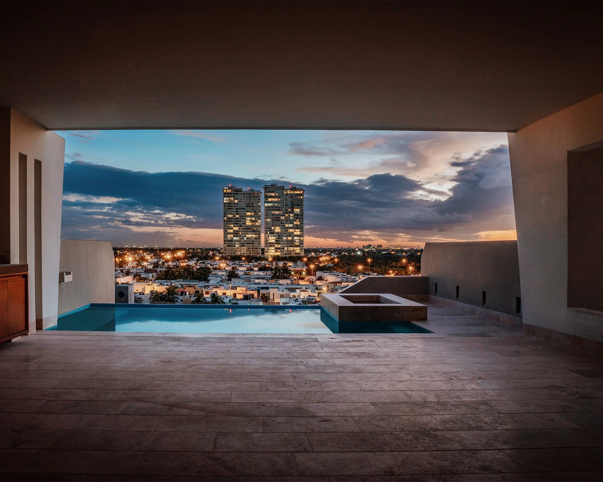 Vista de una piscina en un balcón con pisos de madera, durante el atardecer, con edificios residenciales y un cielo con nubes al fondo.