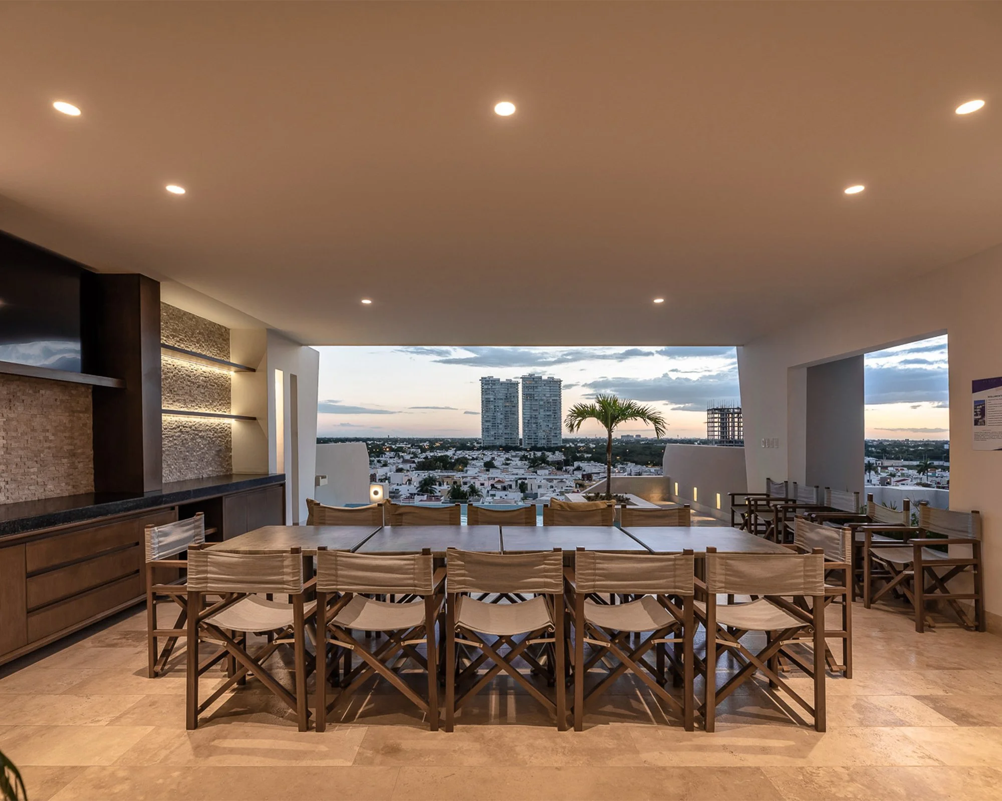 Comedor al aire libre con mesas de madera y sillas, vista del atardecer con edificios altos en el fondo.