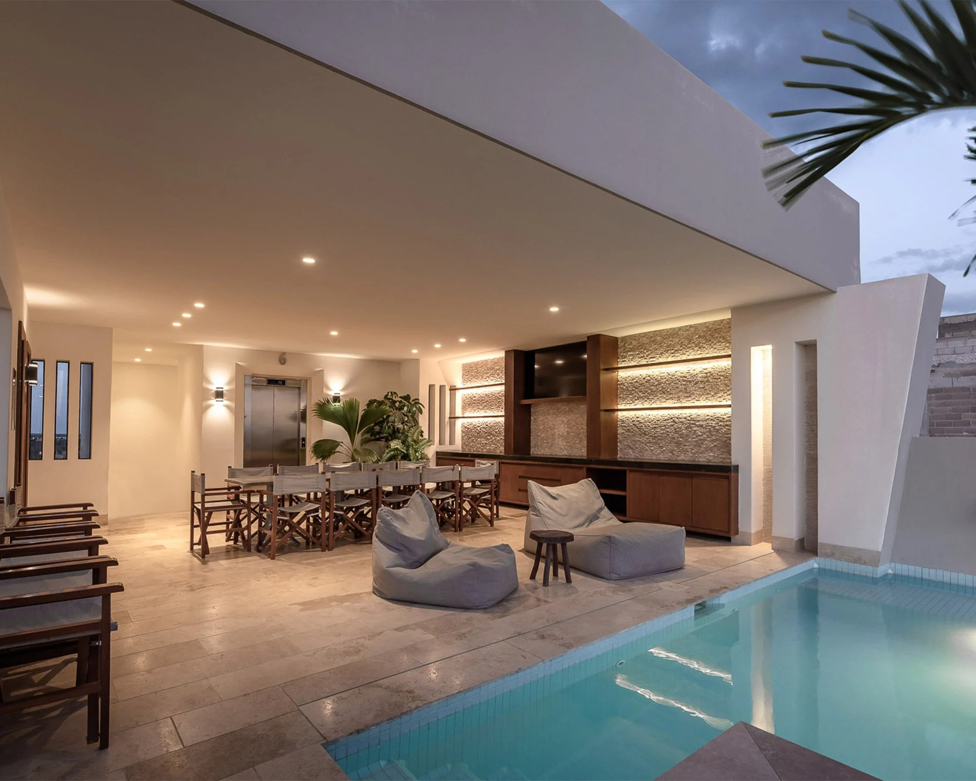 Interior de una casa moderna con piscina, sillas de madera alrededor, áreas de descanso con bancos y cojines, decoraciones con plantas, paredes con texturas y luces cálidas, y ventanas que dejan ver el cielo nocturno.