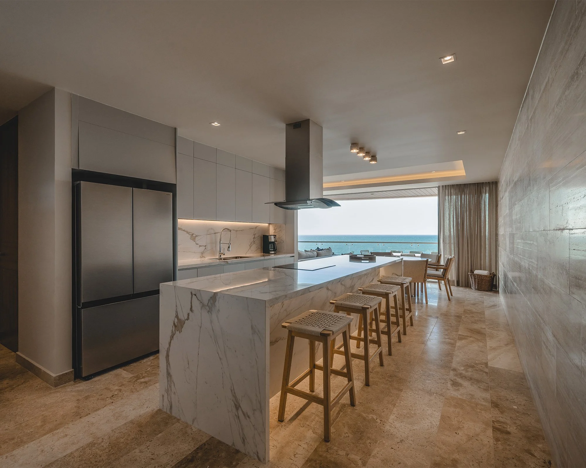 Interiores de una cocina moderna con isla de mármol, sillones de madera, vista al mar y cortinas largas