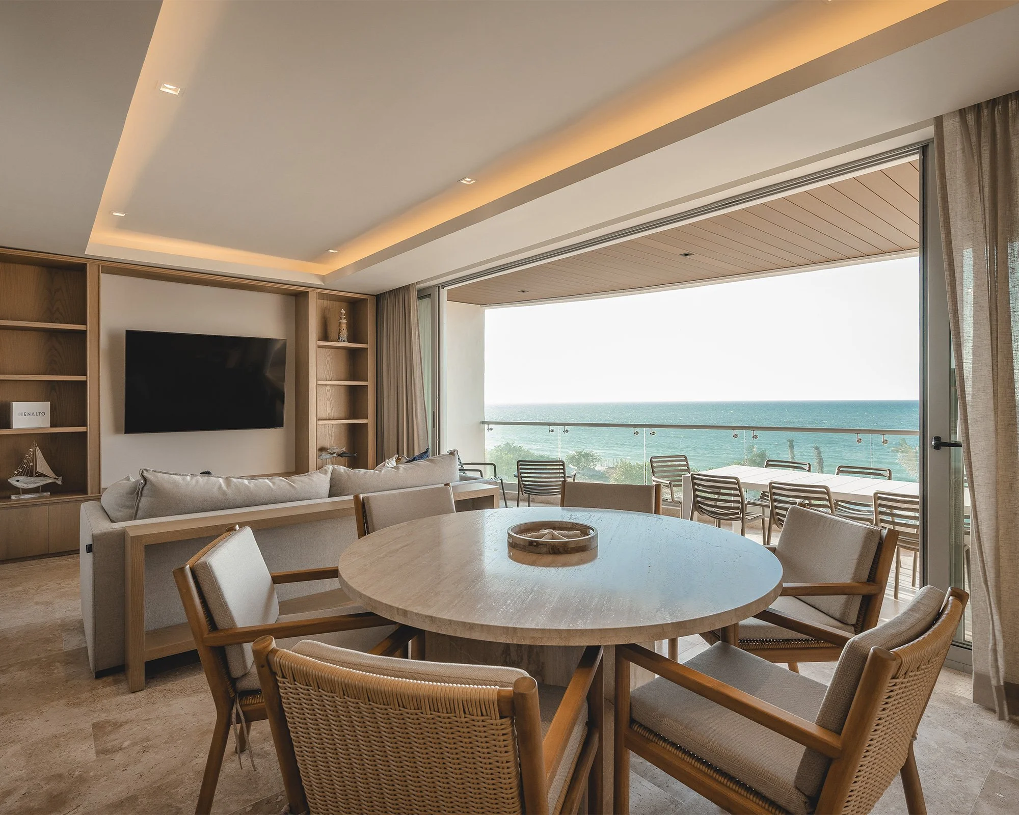 Salón moderno con vista al mar a través de una puerta corrediza, muebles de madera y una mesa redonda en el centro.