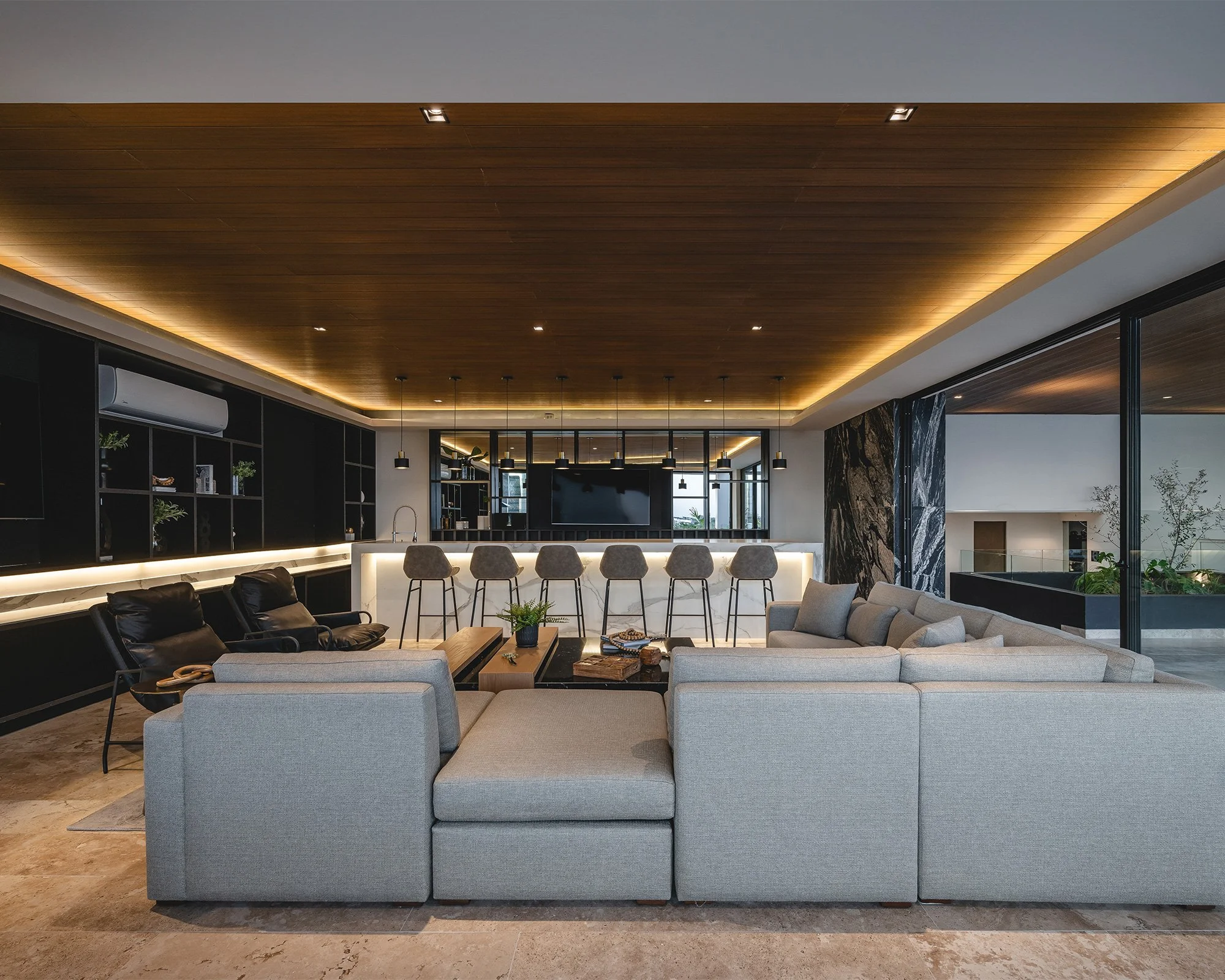 Diseño interior de una sala moderna con muebles cómodos, techo de madera con iluminación integrada y una barra de cocina en el fondo, ventanas grandes que permiten la entrada de luz natural.