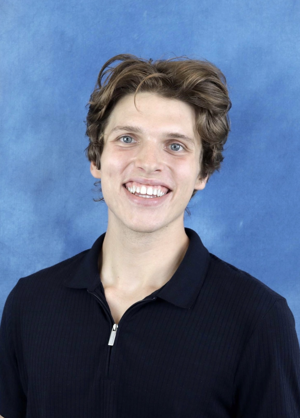 A young man with wavy brown hair, blue eyes, and a big smile, wearing a black zip-up shirt against a blue background.