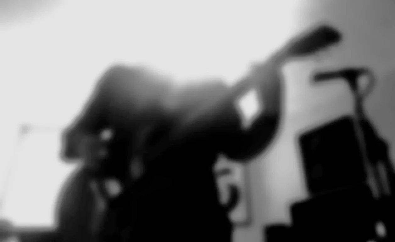 A blurry black-and-white photo of a person with long hair, reaching out with their hand in front of a wall with various boxes or shelves.