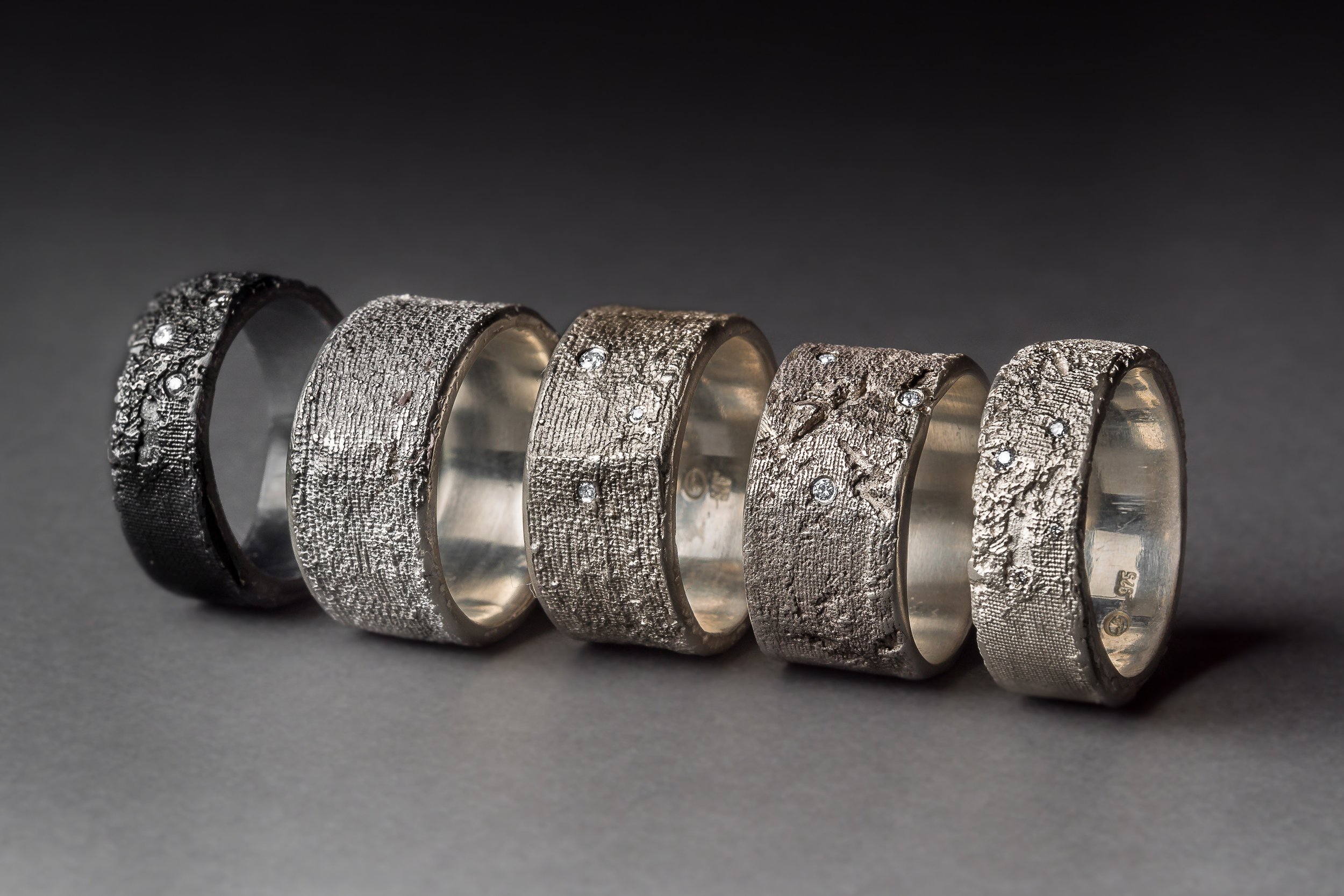 A row of five textured silver rings with embedded diamonds displayed on a dark background.