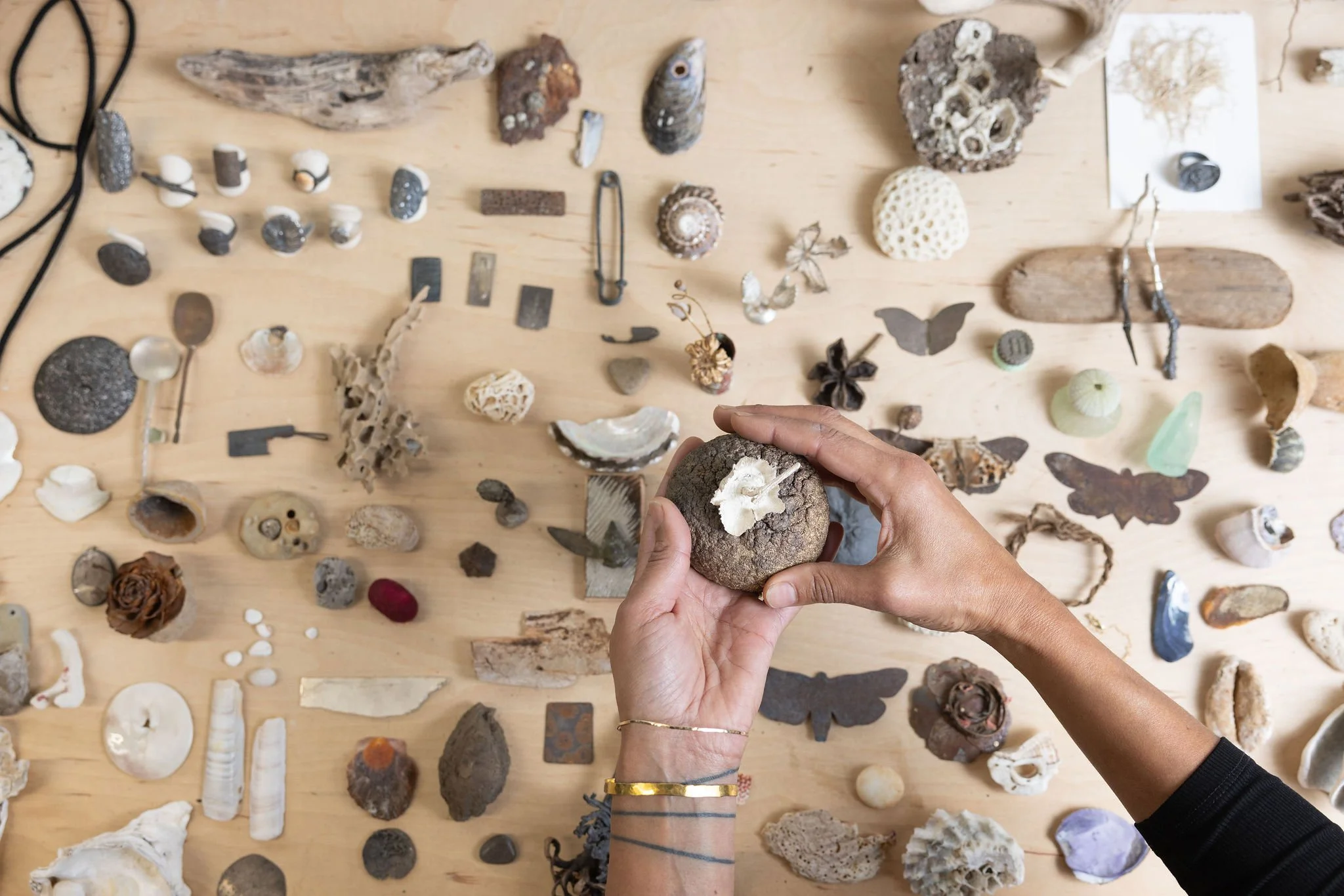 Maya's hands arranging various small rocks, shells, and artifacts on a wooden table. The items include objects from nature and the sea, beads, fossils, and decorative objects.