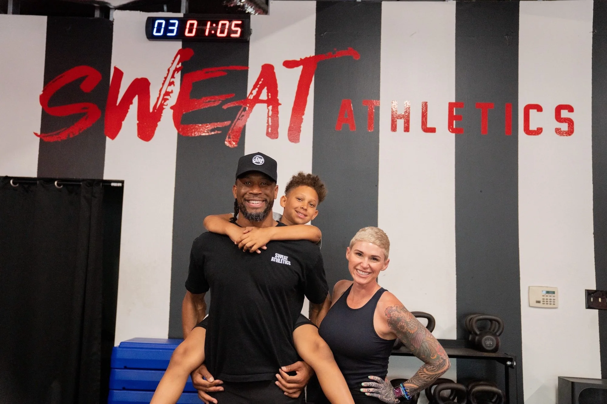 A man and a woman smiling at the camera inside a gym with a large red and black sign that says "Sweat Athletics." The man is holding a young girl with curly hair, and the woman has tattoos on her arm.