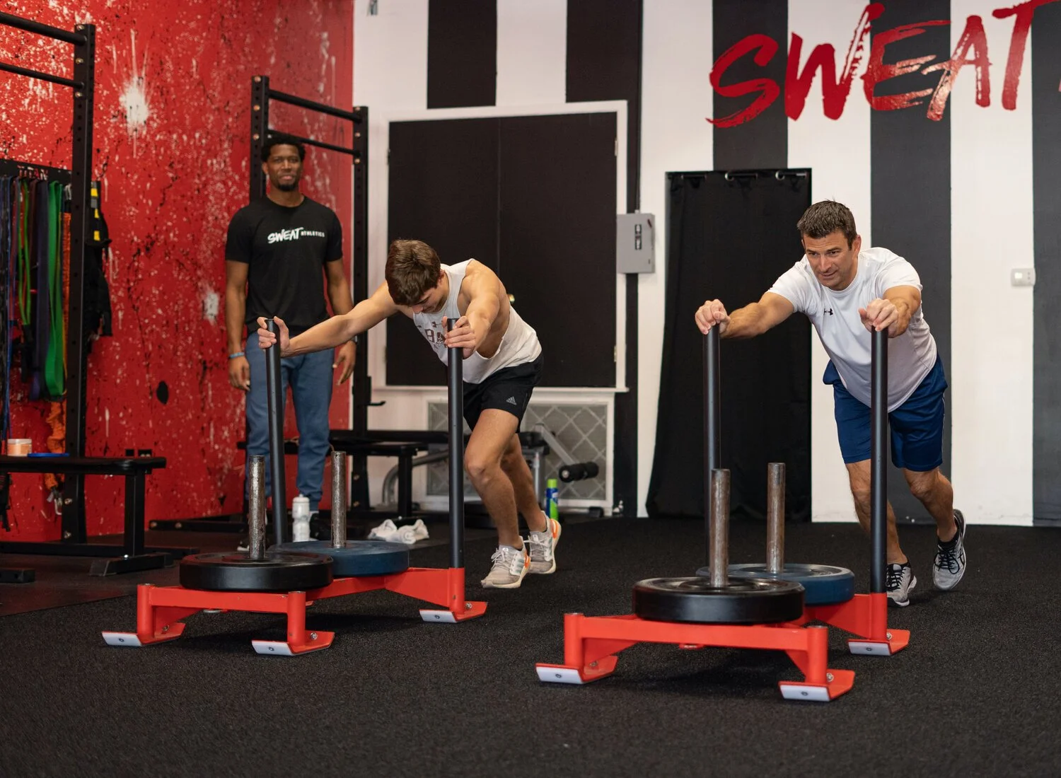 Two men are pushing weighted sleds across a gym floor while a trainer observes. The gym has a red and black color scheme with the word 'SWEAT' written on the wall.