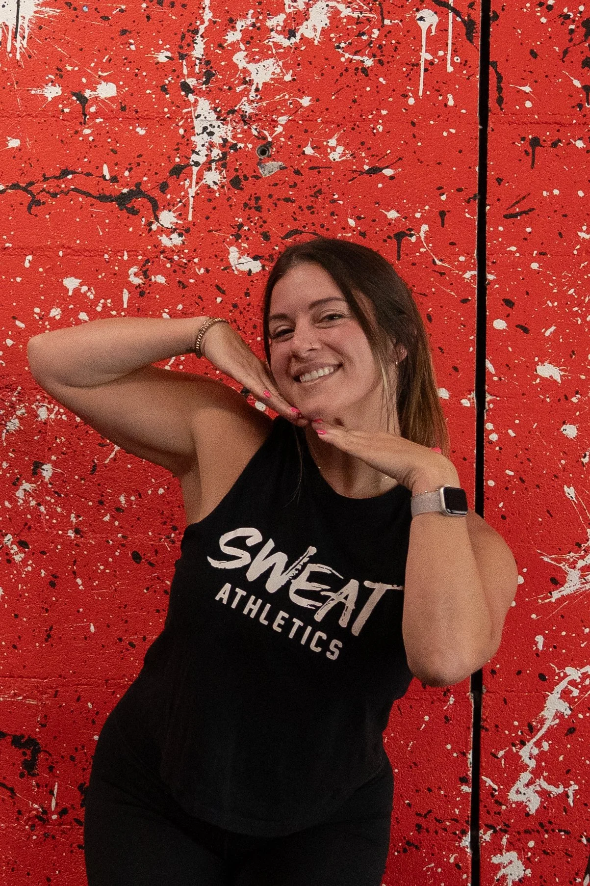 A woman with brown hair smiling and posing in front of a red splattered paint wall. She is wearing a black tank top that says "SWEAT ATHLETICS," a watch on her left wrist, a bracelet on her right wrist, and has pink nail polish.