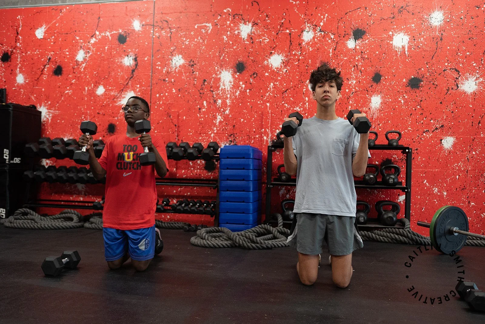 Two young boys kneeling on the floor in a gym, holding dumbbells at shoulder height during a workout session. Behind them, a red wall with black and white splatter paint, racks of weights, and coiled ropes are visible.