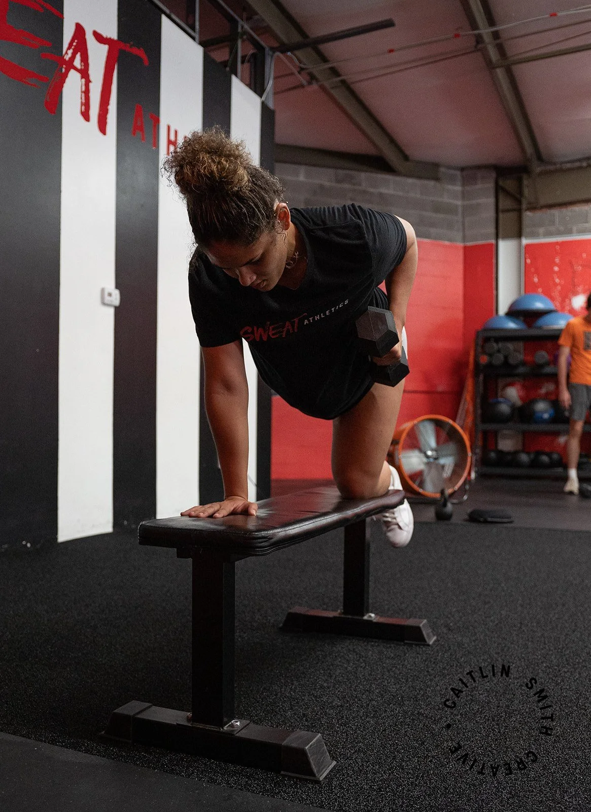 A woman in a black workout shirt with red lettering is doing a knee exercise on a bench inside a gym, holding a dumbbell in her right hand. In the background, there are shelves with medicine balls and a person in an orange shirt.
